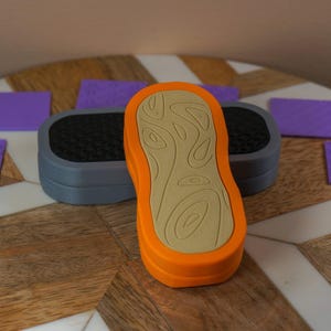 May include: An orange and tan textured shoe sole stamp, with a grey stamp base and several purple textured squares. The items are arranged on a wooden surface with a white geometric pattern.
