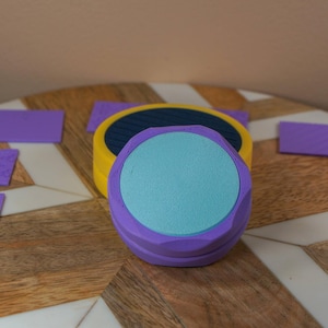 May include: A set of purple, yellow, and blue craft tools and accessories. The image features a round, multi-colored object with a light blue center, surrounded by several small, textured purple rectangles. The items are arranged on a wooden surface.
