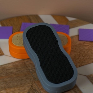 May include: An orange and tan stamp with a black textured base, surrounded by small purple textured stamps. The stamps are on a wooden surface with a white and brown pattern. The base of the stamp is grey.