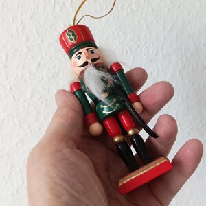 Op de afbeelding: Een kleine notenkraker-ornament met een rode hoed voorzien van een groen hulstblad, een witte baard van imitatiebont en een groen jasje met gouden details. De figuur heeft een rode broek, zwarte laarzen en een rode rechthoekige voet met gouden koord.