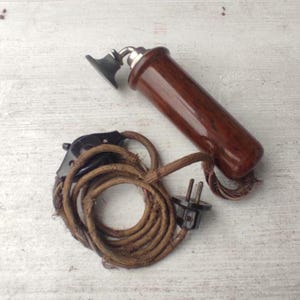 Könnte beinhalten: Ein antiker Telefonhörer mit einem dunkelbraunen Holzgriff und einem schwarzen Ohrhörer. Der Hörer ist mit einem aufgerollten, braunen, stoffummantelten Kabel mit einem schwarzen Stecker verbunden. Ein klassisches Kommunikationsgerät.