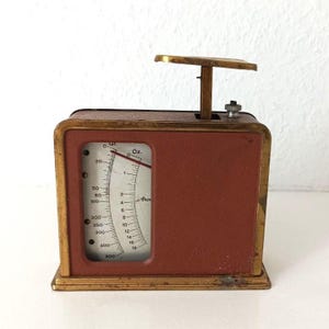 May include: A rectangular mechanical scale with a matte terracotta-colored body and a gold-toned metal frame. The front features a vertical display window with a white dial showing measurements in grams and ounces, with a red needle indicator.