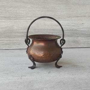 Könnte beinhalten: Ein kleiner, antiker Kupferkessel mit gebogenem Griff und drei dekorativen Füßen. Der Kessel hat eine gehämmerten Textur und eine dunkle, gealterte Patina, was darauf hindeutet, dass er aus der Zeit stammt. Der Griff ist aus dunklem Metall gefertigt, und der Kessel wird wahrscheinlich zu Dekorationszwecken verwendet.