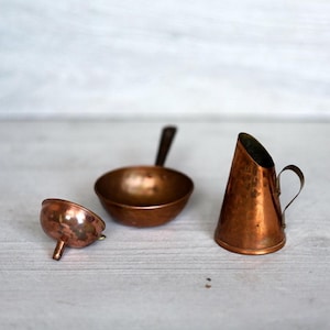 Op de afbeelding: Een verzameling kleine, antieke kookgerei van koper. De set bevat een kleine trechter, een ronde kom met een handvat en een conische kan met een handvat. De items zijn gerangschikt op een lichtgekleurd houten oppervlak.