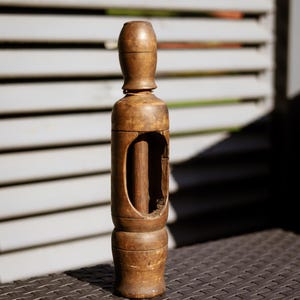 Op de afbeelding: Een cilindervormig bruin houten gereedschap met een afgeronde bovenkant, een verbrede voet en een centrale verticale uitsparing die een dunne interne staaf onthult. Het hout heeft een verweerde, matte afwerking met zichtbare houtnerven.