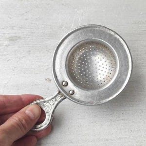 Könnte beinhalten: Ein silberfarbenes Teesieb aus Metall mit einer kreisförmigen Schale und einem perforierten Siebeinsatz in der Mitte. Das Sieb hat einen kurzen, flachen Griff mit einer runden Aussparung am Ende, der mit zwei sichtbaren Nieten befestigt ist.