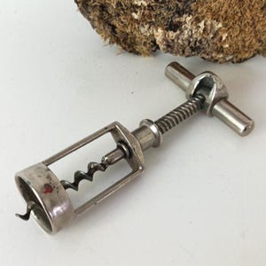 Op de afbeelding: Een vintage zilverkleurige kurkentrekker met een T-vormige handgreep en een spiraalvormige worm. De kurkentrekker heeft een rechthoekig frame en een klein rood merkteken aan de zijkant. De achtergrond bevat een stuk getextureerd, mosachtig hout.