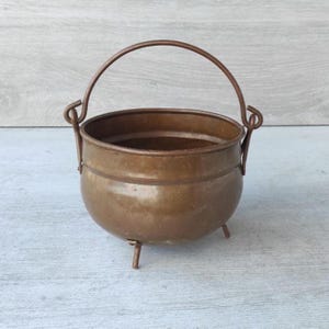 Könnte beinhalten: Ein kleiner, antiker Kupferkessel mit rundem Körper, drei kurzen Beinen und einem gebogenen Griff. Der Kessel hat eine warme, rötlich-braune Farbe und wird wahrscheinlich für dekorative Zwecke oder zum Kochen in kleinem Maßstab verwendet.