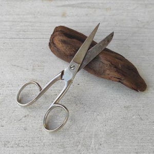 Può includere: Forbici argentate con fori rotondi per le dita, aperte e appoggiate su un pezzo di legno marrone scuro. Le lame sono leggermente aperte, mostrando i bordi affilati. Le forbici sono probabilmente per l'artigianato o il cucito.
