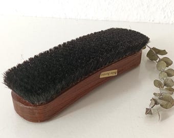 May include: A wooden shoe brush with dark bristles. The brush has a curved, rectangular shape with a brown wooden handle. The text "Reine Borsten" is visible on a small label on the side of the brush. A sprig of dried leaves is next to the brush.