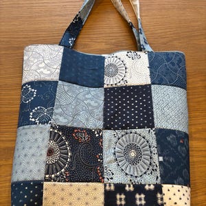 May include: A patchwork tote bag featuring a variety of blue and white patterned squares. The bag has two handles and a zip closure. The patterns include floral, geometric, and dotted designs. The bag is made of fabric.
