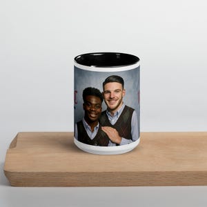 May include: White ceramic mug with a black rim. The mug features a photo of two men, one Black and one White, smiling and wearing sweaters. The mug has a black background and the text "It's Not Magic, It's Love" in red.