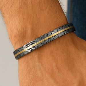 May include: A patterned black and silver bracelet with a gold stripe is displayed on a wrist. The cuff bracelet features a central gold-coloured stripe. The bracelet is a stylish accessory.