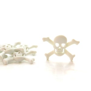 Puede incluir: Un montón de dijes de plástico blanco en forma de calavera y huesos cruzados, con un dije más grande de calavera y huesos cruzados a la derecha.