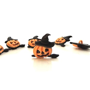 May include: A collection of Halloween-themed decorative buttons. Each button features a carved orange pumpkin wearing a black witch hat. The pumpkins have carved faces and are lying on a white surface. Some have a black broom.