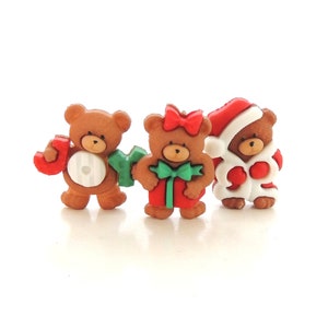 May include: Three brown teddy bear buttons with Christmas themes. One bear is wearing a Santa hat, one is holding a green and red present, and one is holding a green and red "JOY" sign.