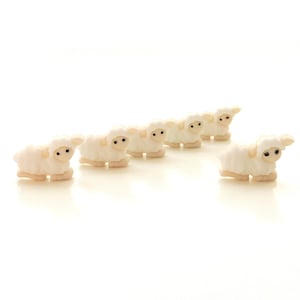 May include: A line of six white ceramic sheep figurines with black eyes, standing in a row on a white background.