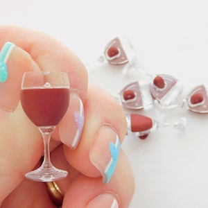 May include: A miniature glass of red wine held in a hand with blue and white nail polish. The glass is clear and the wine is a deep red color.