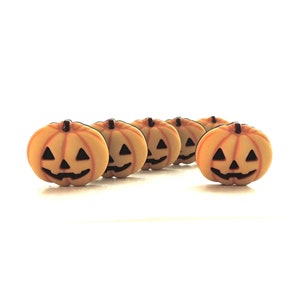 May include: A set of five small, orange pumpkin-shaped buttons, each featuring a carved jack-o'-lantern face with black triangular eyes, a nose, and a jagged mouth. The buttons have a smooth, rounded surface and a dark brown stem at the top.