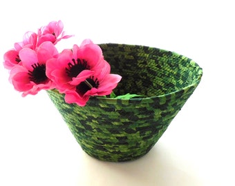 Puede incluir: Una cesta tejida con un patrón de camuflaje verde y negro, que contiene varias flores rosas brillantes con centros negros. La cesta tiene forma cónica y está sobre una superficie blanca. Las flores están agrupadas.