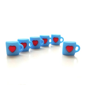 May include: Six small, light blue mugs with red heart designs. Each mug has a handle and is arranged on a white surface. The mugs are evenly spaced, with one slightly offset from the others. The overall aesthetic is simple and charming.