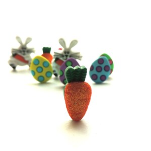 May include: A collection of Easter-themed decorations, including bunnies, eggs, and a carrot. The carrot is orange with green leaves and a sparkly texture. The eggs are decorated with colorful polka dots and stripes. The bunnies are white with pink ears and noses.