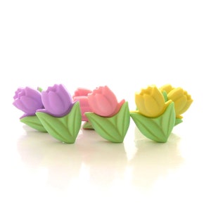 May include: A collection of small, tulip-shaped buttons in pastel colors including purple, pink, and yellow. Each button features a distinct green leaf base and a rounded flower head, arranged in a horizontal row against a plain white background.