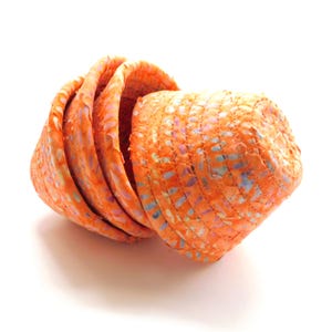 May include: An orange woven basket with a textured surface. The basket has a conical shape and is made of a material with flecks of blue, purple, and yellow. The basket is partially nested.