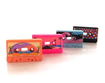 May include: Four rectangular cassette tape shaped pins featuring distinct designs. The orange pin shows a sunset beach scene, the pink pin depicts a palm tree road, the light blue pin displays a night sky, and the black pin has a diamond pattern.