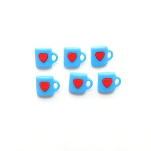 May include: Six small, light blue mug-shaped embellishments, each with a red heart in the center. The mugs have a handle on the right side. The embellishments are arranged on a white background.