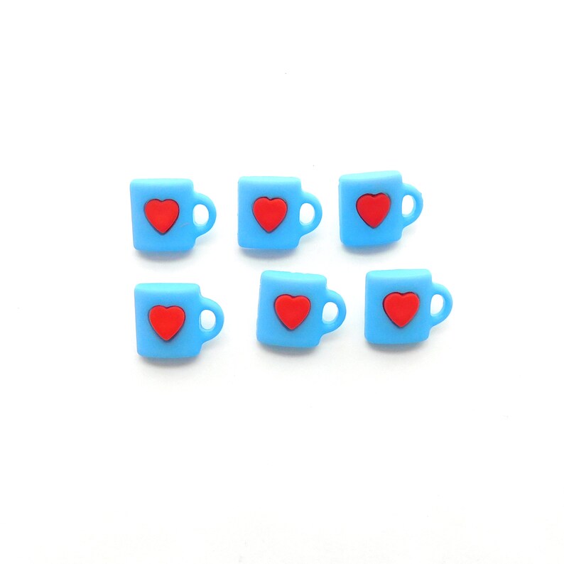 May include: Six small, light blue mug-shaped embellishments, each with a red heart in the center. The mugs have a handle on the right side. The embellishments are arranged on a white background.