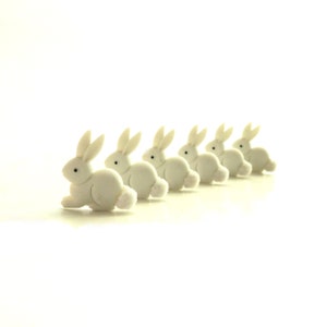 May include: A line of six white ceramic bunny figurines, each with a fluffy tail, arranged in a row on a white background.