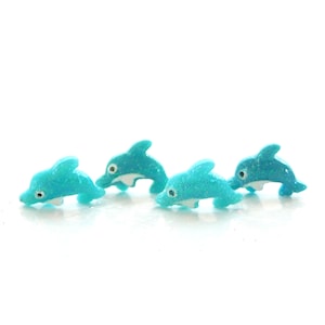 May include: Four turquoise blue glitter dolphin figurines. The dolphins are in a swimming position with their tails up and their heads down.