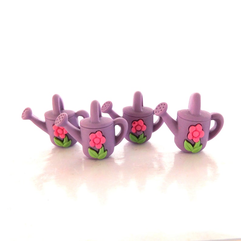 May include: Four small, lavender watering cans with pink flower and green leaf accents. Each miniature watering can has a handle, a spout, and a decorative flower. The watering cans are arranged in a row on a white surface.