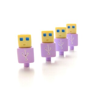 May include: Four USB connectors with a cartoon face design. The top of the connector is yellow with blue eyes and a small mouth. The base is lavender with a USB symbol. The connectors are arranged in a row against a white background.