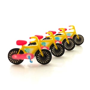 Puede incluir: Una fila de cuatro bicicletas de juguete en miniatura de color amarillo, rosa y azul con ruedas negras. Las bicicletas están alineadas en una fila, con la rueda delantera de cada bicicleta tocando la rueda trasera de la bicicleta que está delante.