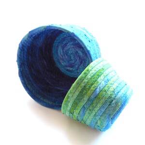 May include: Two handmade, round baskets. One is a deep blue with a spiral design inside. The other is smaller, with a green and blue striped pattern. Both are made of a woven material.