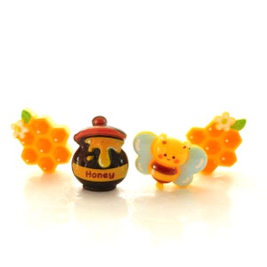 May include: A collection of bee-themed charms. Includes a honey pot with the word "Honey", a cartoon bee with blue wings, and two honeycomb shapes with white flowers and green leaves. The charms are yellow, brown, and orange.