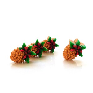May include: Four decorative pinecone ornaments. The pinecones are a warm brown color, with green holly leaves and red bows. The ornaments are arranged on a white surface.