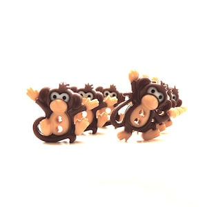 Puede incluir: Una colección de botones marrones con forma de mono y caras y manos color beige. Los botones están dispuestos en semicírculo, con algunos monos levantando los brazos. Los botones tienen dos o tres agujeros para coser.