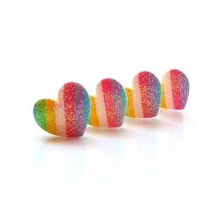 Puede incluir: Cuatro decoraciones en forma de corazón, brillantes, con rayas de arcoíris. Los corazones están dispuestos en una fila, con las puntas de los corazones apuntando hacia la derecha.