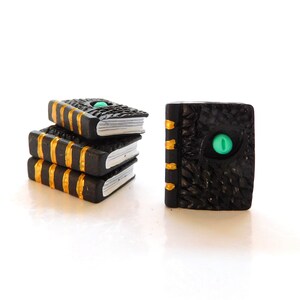 May include: A stack of three black books with gold trim and a single black book with a green eye on the cover. The books are made of polymer clay and are miniature in size.