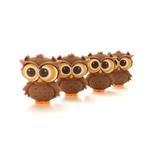May include: Four brown owl figurines with large, round eyes and orange feet are arranged in a row. The owls have a cartoonish design, with a light brown face and a darker brown body. The background is plain white.