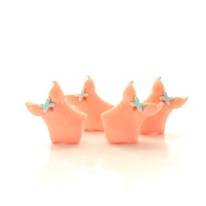 May include: Four pink ceramic pig figurines with blue cross details. The pigs are facing forward with their heads tilted slightly down.