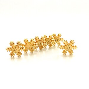 May include: A collection of six gold-toned snowflake-shaped charms arranged in a horizontal row. Each charm features a detailed, symmetrical six-pointed star design with intricate cutouts and a polished metallic finish throughout.