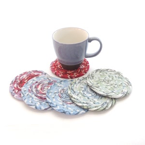 May include: Five colorful fabric coasters with a blue mug on top of a red coaster. The coasters are made of woven fabric and have a circular shape.