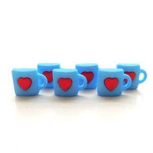 May include: Six light blue mugs with red heart designs. Each mug has a handle and a heart-shaped accent on the front. The mugs are arranged on a white surface.