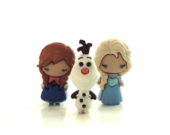 May include: Three small decorative buttons shaped like cartoon characters. On the left, a button with brown hair and a blue dress. In the center, a white snowman button with a carrot nose and brown twigs. On the right, a button with blonde hair and a light blue dress.