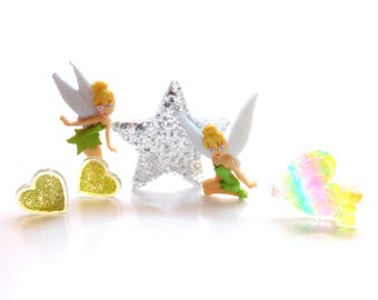 May include: Two small fairy figurines with blonde hair, white wings, and green dresses, accompanied by a silver glitter star, two gold glitter heart-shaped charms, and one iridescent, translucent heart-shaped charm on a white background.