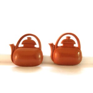 Puede incluir: Dos pendientes de cerámica marrón en forma de tetera. Las teteras tienen un cuerpo redondeado y una pequeña tapa en la parte superior.